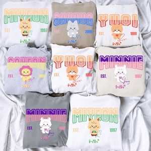 Peut inclure: Une collection de huit sweats à capuche présentant des graphismes de personnages de dessins animés, des noms comme MIYEON, SHUHUA, YUQI, SOYEON et MINNIE, des années de naissance et des signatures. Les sweats sont de diverses couleurs.