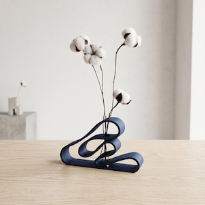 May include: A contemporary navy blue vase with an abstract, ribbon-like design. The vase contains cotton flowers with white, fluffy heads and brown stems. The vase is placed on a light-coloured wooden surface.