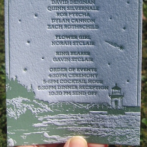 May include: A light blue wedding invitation card with a white embossed design. The card features the names Tessa & Charlie, the wedding date, and a list of the wedding party members. The card also includes a small illustration of a lighthouse and a seascape.