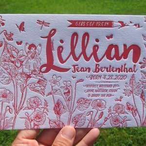 May include: A red and white birth announcement card with the name "Lillian" in large script. The card includes the baby's weight, length, and birth date. Floral and fairy illustrations surround the text.