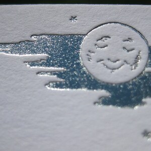 May include: A silver embossed image of a sleeping moon with a face and a cloud behind it. The image is on a white background.