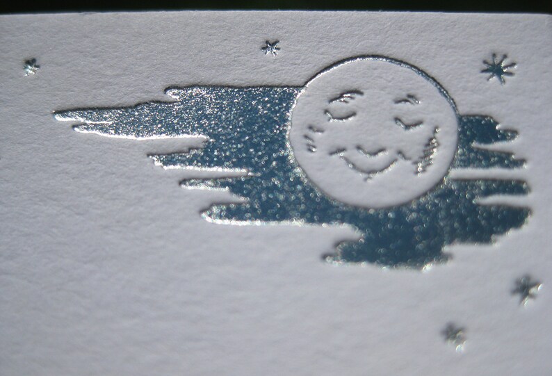 May include: A silver embossed image of a sleeping moon with a face and a cloud behind it. The image is on a white background.