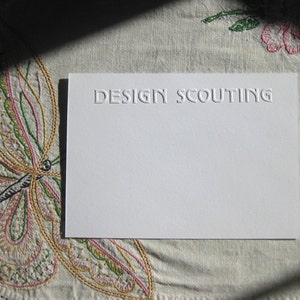 May include: A white card with the text "DESIGN SCOUTING" embossed in a dark gray color. The card is lying on a white fabric with an embroidered butterfly design.