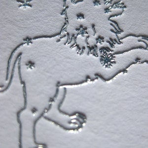 May include: A silver embossed image of the constellation Leo, the lion, with stars surrounding the lion.