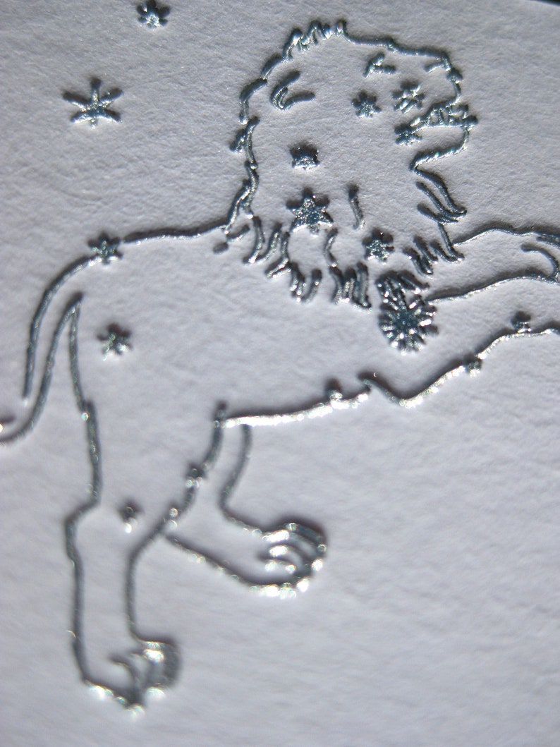 May include: A silver embossed image of the constellation Leo, the lion, with stars surrounding the lion.
