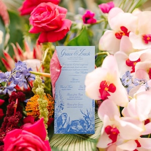 May include: A wedding invitation with the names "Grace & Zack" is surrounded by vibrant flowers. The invitation is light blue with blue illustrations and text. Red roses, white orchids with red centers, and other colorful blooms create a festive backdrop.