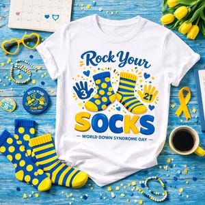 Peut inclure: T-shirt blanc avec l'inscription "Rock Your Socks" et un motif de chaussettes bleues et jaunes. Le t-shirt est entouré de chaussettes, d'un calendrier et d'autres accessoires sur une surface en bois bleu. Le texte "World Down Syndrome Day" est également présent.