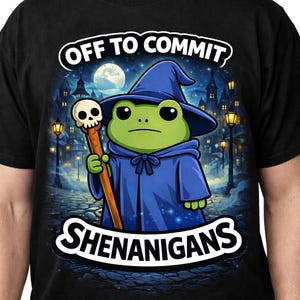 May include: Black t-shirt featuring a cartoon frog dressed as a wizard, holding a skull-topped staff. The frog wears a blue robe and hat, with the words "OFF TO COMMIT SHENANIGANS" in a white, stylized font.