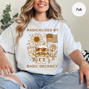 Könnte beinhalten: Aschgraues T-Shirt mit brauner Grafik. Die Grafik zeigt einen Burger mit Sonnenbrille, der ein Getränk und eine amerikanische Flagge hält, mit einer Sonnenblume und einer Rose. Der Text lautet "RADICALIZED BY BASIC DECENCY."