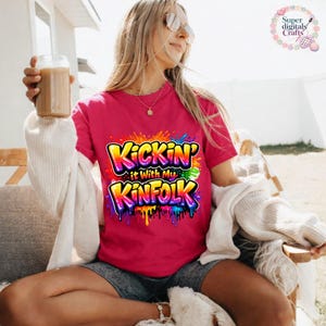 May include: A magenta t-shirt with the text "Kickin' it With My Kinfolk" in a colorful graffiti-style design. The graphic features yellow, orange, blue, and green colors with dripping paint effects. The shirt is short-sleeved.