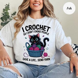 May include: Ash-colored t-shirt with the text "I CROCHET SO I DON'T CHOKE PEOPLE" and "SAVE A LIFE... SEND YARN." A black cat sits in a blue basket filled with yarn balls. The cat has blue eyes and is surrounded by yarn.