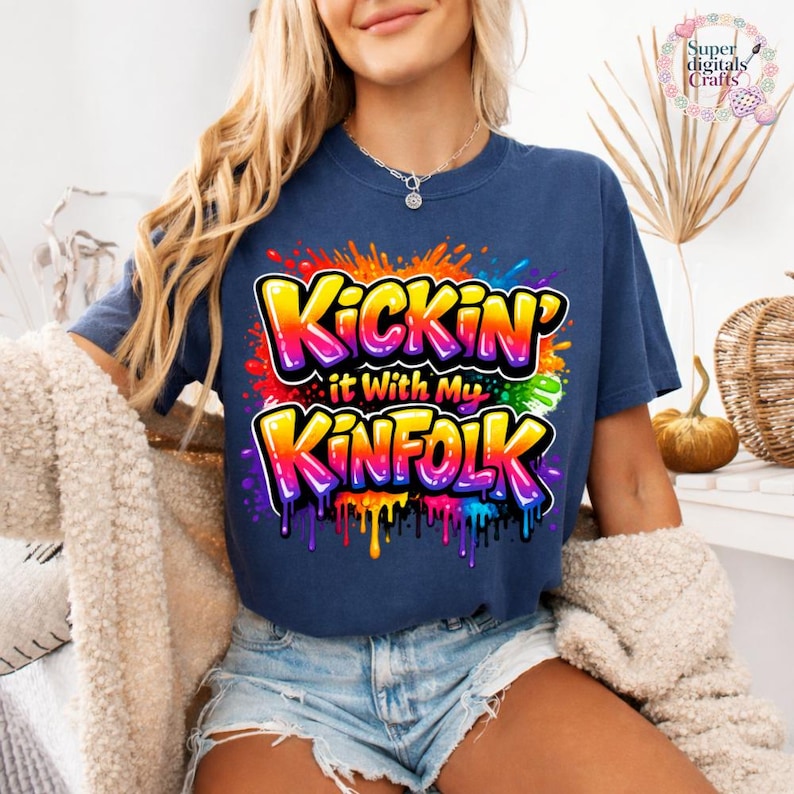 May include: A navy blue t-shirt with the text "Kickin' it With My Kinfolk" in a colorful graffiti-style design. The text is outlined in yellow and features a rainbow gradient. The shirt is worn with denim shorts.