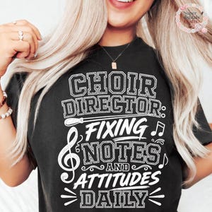 Puede incluir: Una camiseta negra de manga corta con texto blanco que dice "CHOIR DIRECTOR: FIXING NOTES AND ATTITUDES DAILY" junto con un gráfico de una batuta de director, una clave de sol y notas musicales en un diseño apilado.