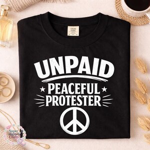 Pode incluir: Camiseta preta com a frase branca "UNPAID PEACEFUL PROTESTER" e um s&iacute;mbolo da paz. A camiseta est&aacute; dobrada. Acess&oacute;rios, incluindo uma x&iacute;cara e joias, s&atilde;o vis&iacute;veis no fundo.