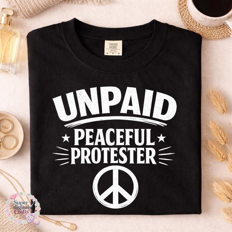 Pode incluir: Camiseta preta com a frase branca "UNPAID PEACEFUL PROTESTER" e um s&iacute;mbolo da paz. A camiseta est&aacute; dobrada. Acess&oacute;rios, incluindo uma x&iacute;cara e joias, s&atilde;o vis&iacute;veis no fundo.