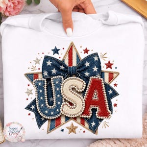 Op de afbeelding: Witte sweatshirt met een patriottisch "USA" ontwerp in rood, wit en blauw. Het ontwerp bevat een denim-achtige strik, sterren en een sterrenvorm, perfect voor het vieren van Amerikaanse feestdagen.