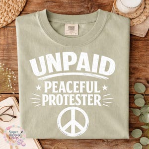 Pode incluir: Camiseta verde s&aacute;lvia com as palavras brancas "UNPAID PEACEFUL PROTESTER" e um s&iacute;mbolo de paz. A camiseta tem gola redonda e uma etiqueta Comfort Colors. O design est&aacute; centralizado na frente.