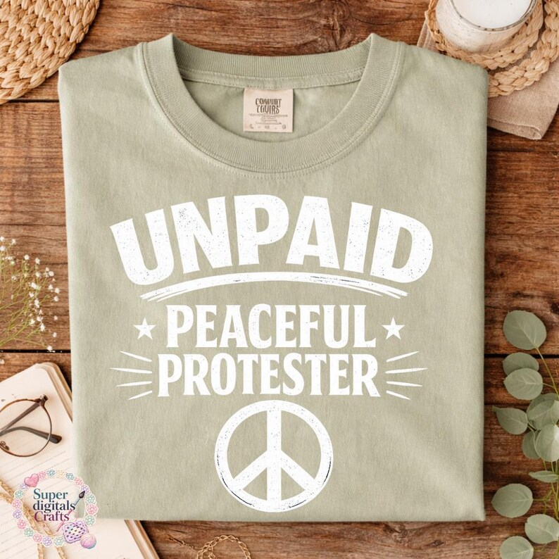 Pode incluir: Camiseta verde s&aacute;lvia com as palavras brancas "UNPAID PEACEFUL PROTESTER" e um s&iacute;mbolo de paz. A camiseta tem gola redonda e uma etiqueta Comfort Colors. O design est&aacute; centralizado na frente.