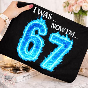 May include: Black pillowcase with the text "I WAS... NOW I'M..." in white. The number "67" is displayed in bright blue flames. The pillowcase is held up by a person.