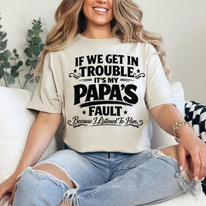 May include: Beige t-shirt with the text "IF WE GET IN TROUBLE IT'S MY PAPA'S FAULT Because I Listened To Him." in black. The text is arranged in a playful, bold font with star accents. The shirt is worn by a person.