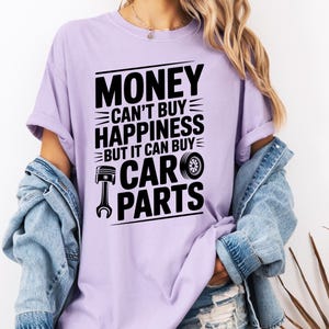 May include: A light purple t-shirt with the text "MONEY CAN'T BUY HAPPINESS BUT IT CAN BUY CAR PARTS" in black. The design includes a piston and a tire. The person is wearing a denim jacket.