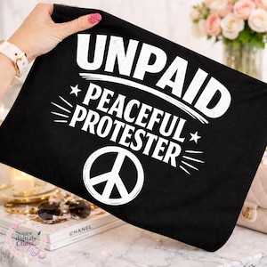 Pode incluir: Camiseta preta com a frase branca "UNPAID PEACEFUL PROTESTER" e um s&iacute;mbolo da paz. A camiseta &eacute; segurada por uma pessoa. O fundo inclui um livro, &oacute;culos de sol e flores.