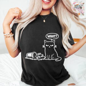 May include: Black t-shirt featuring a white graphic of a cat with an angry expression and the word "WHAT?" in a speech bubble. The cat is next to a tipped-over plant pot. The shirt is a casual style.