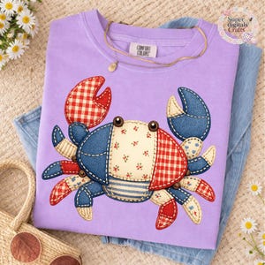 May include: A lilac Comfort Colors t-shirt featuring a crab applique. The crab design incorporates red and white gingham, blue denim, and floral fabric. A gold necklace with a pendant rests on the shirt. The shirt is folded on top of a pair of blue jeans.
