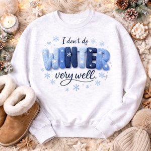 May include: White sweatshirt with the text "I don't do WINTER very well" in blue and white. The word WINTER is in a 3D knit style. Blue snowflakes are scattered around the text.