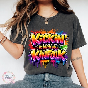 May include: Dark gray t-shirt with the text "Kickin' it With My Kinfolk" in a colorful, graffiti-style design. The text is outlined in white with a vibrant rainbow gradient. The shirt has a crew neck and short sleeves.
