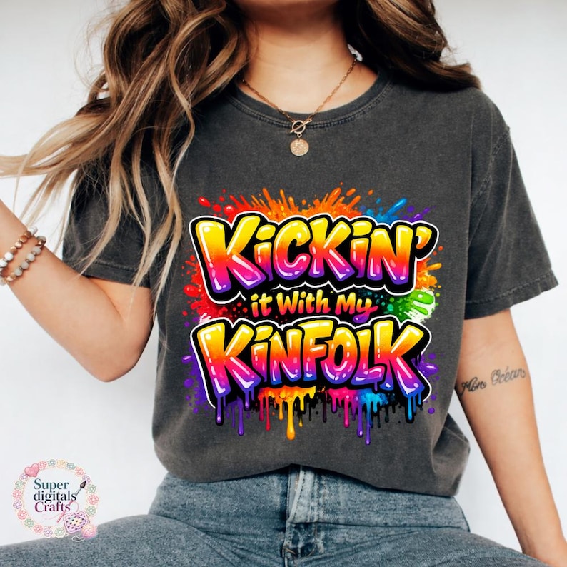 May include: Dark gray t-shirt with the text "Kickin' it With My Kinfolk" in a colorful, graffiti-style design. The text is outlined in white with a vibrant rainbow gradient. The shirt has a crew neck and short sleeves.