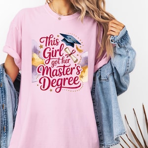 Può includere: Una maglietta rosa chiaro con la scritta "This Girl got her Master's Degree" in corsivo. Il design include un cappello di laurea, un diploma e stelle dorate. La maglietta è in stile casual.