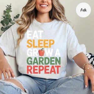 May include: Light gray t-shirt with the words "EAT SLEEP GROW A GARDEN REPEAT" in white, orange, green, and red. A red pepper graphic replaces the letter "O" in "GROW". The shirt is a casual, short-sleeved style.