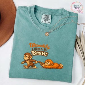 Op de afbeelding: Een Comfort Colors t-shirt in zeegroen met de tekst "Ultimately I Have to Be Brave" en een cartoon aap ontwerp. Een zilveren ketting met hanger ligt op het shirt. Maat S-CH.