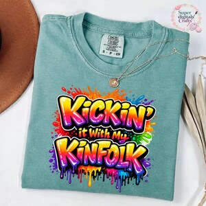 May include: A teal Comfort Colors t-shirt with the text "Kickin' it With My Kinfolk" in a colorful graffiti-style design. The shirt has a crew neck and short sleeves. A silver necklace with a pendant is draped on the shirt.