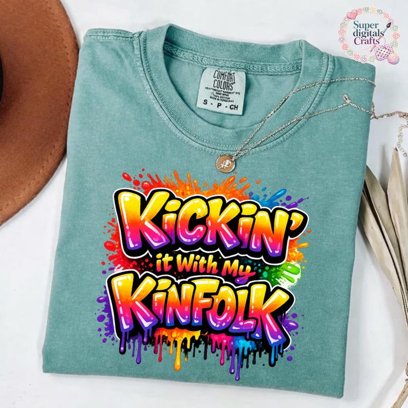 May include: A teal Comfort Colors t-shirt with the text "Kickin' it With My Kinfolk" in a colorful graffiti-style design. The shirt has a crew neck and short sleeves. A silver necklace with a pendant is draped on the shirt.