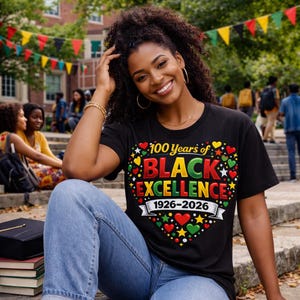 May include: Black t-shirt with a heart-shaped design that reads "100 Years of BLACK EXCELLENCE 1926-2026" in red, green, yellow, and black. The shirt is worn by a person sitting outdoors.