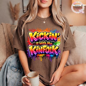 May include: A brown t-shirt with the text "Kickin' it With My Kinfold" in a colorful, graffiti-style design. The graphic features vibrant rainbow colors with dripping paint effects. The shirt is a casual, comfortable style.