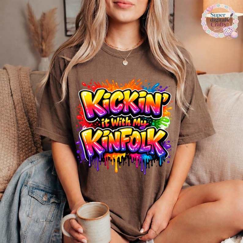 May include: A brown t-shirt with the text "Kickin' it With My Kinfold" in a colorful, graffiti-style design. The graphic features vibrant rainbow colors with dripping paint effects. The shirt is a casual, comfortable style.