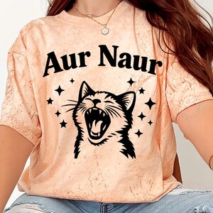 Peut inclure: T-shirt pêche avec l'inscription noire "Aur Naur", au-dessus d'une illustration en noir et blanc d'un chat gueule ouverte. Le motif comprend des étoiles noires autour du chat.