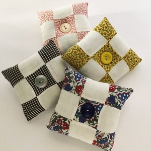 Puede incluir: Cuatro alfileteros de tela con diferentes patrones y colores. Cada alfiletero tiene un botón en el centro. Los patrones incluyen diseños a cuadros, florales y geométricos. Los colores incluyen blanco, negro, marrón, amarillo, rojo, azul y verde.