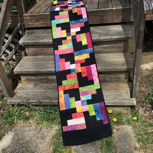 Puede incluir: Camino de mesa rectangular con fondo negro y diseño patchwork. El camino de mesa presenta bloques de colores brillantes, incluyendo rosa, verde, azul y naranja. Los bordes están acabados con un borde negro y costuras decorativas.