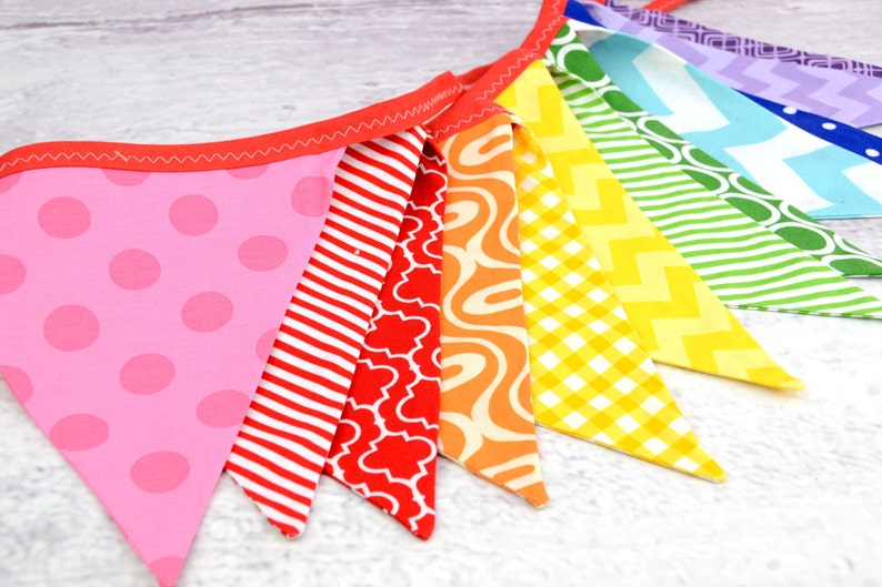 May include: A rainbow-coloured fabric banner with triangular flags. The flags are sewn together and feature different patterns, including polka dots, stripes, swirls, and geometric designs.
