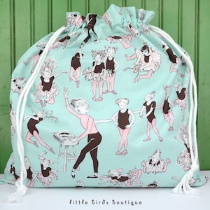 May include: A light blue drawstring bag with a white drawstring. The bag is printed with a pattern of ballerinas in various poses. The text "little birds boutique" is printed on the bottom of the bag.