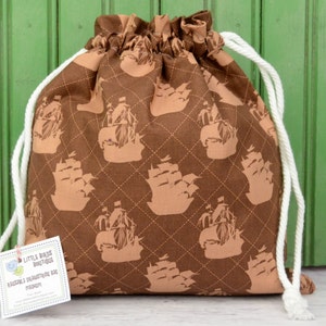 May include: Brown drawstring bag with a repeating pattern of tan sailing ships. The bag has a white drawstring closure and a tag that reads "Little Birds Boutique". The bag is made of fabric and has a diamond pattern.