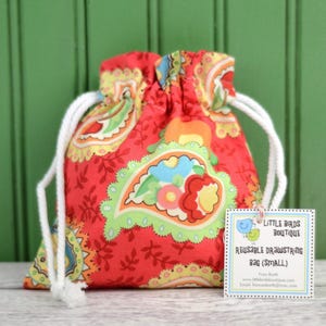 May include: A red drawstring bag with a paisley pattern in shades of green, yellow, blue, and orange. The bag has a white drawstring and a tag that reads "Little Birds Boutique Reusable Drawstring Bag (Small)".