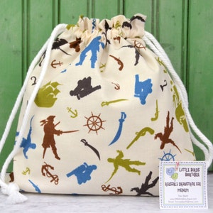 May include: A white drawstring bag with a pirate theme print. The bag features silhouettes of pirates, ships, anchors, swords, and cannons. The bag is made of cotton fabric and has a white drawstring closure. The bag is sitting on a white surface with a tag that reads "Little Birds Boutique Reusable Drawstring Bag Medium".