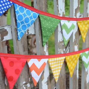 May include: A colourful fabric banner with red trim, featuring various patterns including polka dots, stripes and zigzags. The banner is strung on a wooden fence.