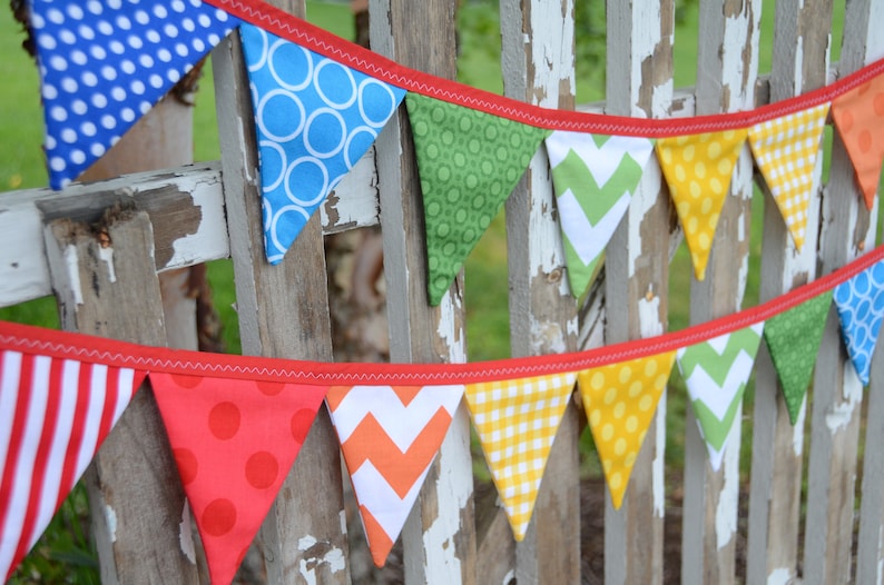 May include: A colourful fabric banner with red trim, featuring various patterns including polka dots, stripes and zigzags. The banner is strung on a wooden fence.