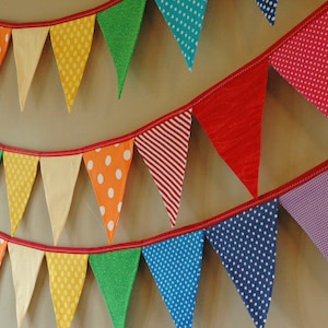 May include: A vibrant bunting banner featuring triangular flags in a variety of patterns and colours, such as red, orange, yellow, green, blue, and purple. The flags are strung on a red ribbon, creating a cheerful decoration.